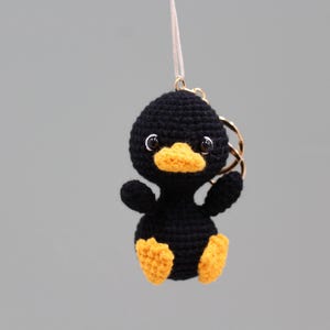 May include: A small, black, crocheted duck keychain featuring a bright yellow beak and matching yellow feet. The duck has two black, glossy eyes and is attached to a gold-toned metal keyring by a thin, white string, hanging against a neutral background.