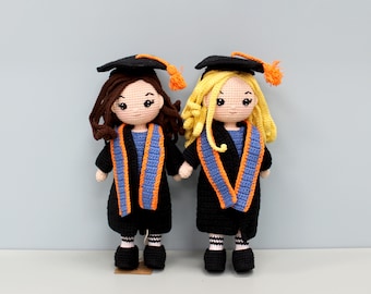 May include: Two handmade crochet dolls dressed in graduation attire. Each doll wears a black cap with an orange tassel, a black gown, and a blue and orange sash. One doll has brown hair, and the other has blonde hair. The dolls are standing side by side.