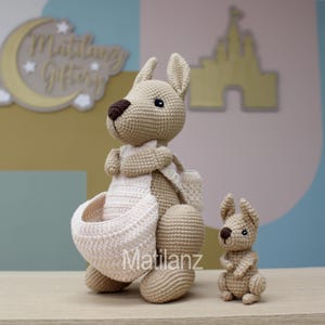 May include: Two crocheted kangaroo plushies, one large and one small. The larger kangaroo is beige with a white pouch and brown eyes. The smaller kangaroo is beige with brown eyes. Both kangaroos are standing on a wooden surface.