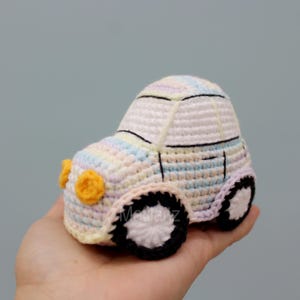 Handmade crocheted car toy in pastel colors with a white body, colorful stripes, yellow headlights, black wheels, and detailed stitching.