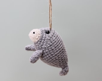 May include: A small, gray, crocheted manatee ornament with a light beige snout and a single black bead eye. The plush figure is suspended by a thin, brown jute twine string attached to the top, featuring a rounded body and small flippers.