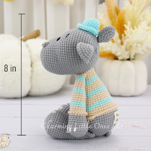 Handmade gray crocheted hippo toy wearing a blue hat and striped scarf, sitting upright at approximately 8 inches tall.