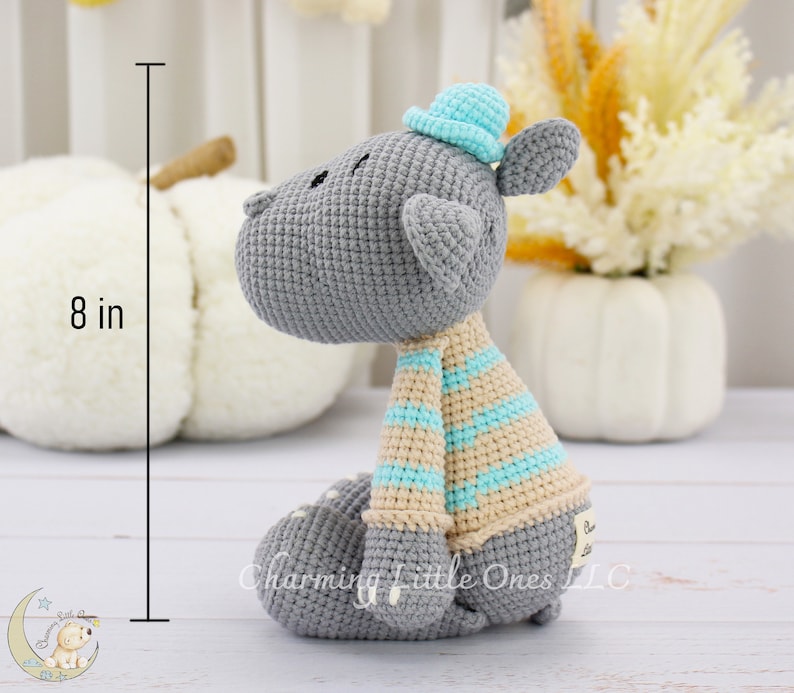 Handmade gray crocheted hippo toy wearing a blue hat and striped scarf, sitting upright at approximately 8 inches tall.