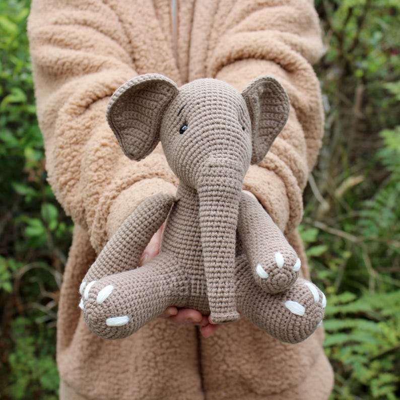 A soft, crocheted elephant toy in shades of brown, featuring oversized floppy ears, a textured trunk threaded with beige yarn, and white painted toes.
