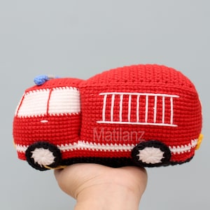 Handmade crocheted fire truck toy in vibrant red with white accents, black wheels, yellow details, and a blue roof, crafted from soft yarn for a cozy texture.