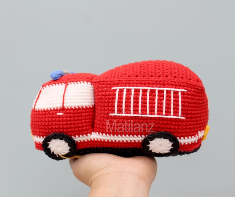 Handmade crocheted fire truck toy in vibrant red with white accents, black wheels, yellow details, and a blue roof, crafted from soft yarn for a cozy texture.