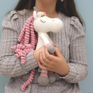 Könnte beinhalten: Eine handgefertigte Häkel-Einhorn-Puppe mit weißem Körper, rosa Mähne und grauen Hufen. Das Einhorn hat einen Blumenkranz und geschlossene Augen. Die Puppe wird vor einem hellblauen Hintergrund gehalten.