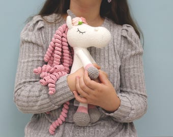 May include: A handmade crochet unicorn doll with a white body, pink mane, and gray hooves. The unicorn has a flower crown and closed eyes. The doll is held against a light blue background.