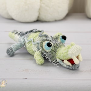 May include: A crocheted alligator toy with green, gray, and blue stripes. The alligator has large, blue eyes and a toothy grin.