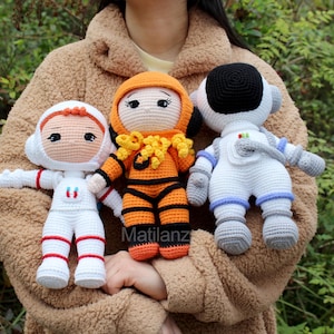 May include: Three crocheted astronaut dolls. One doll is wearing an orange and yellow spacesuit, one is wearing a white spacesuit with blue accents, and one is wearing a white spacesuit with red accents.