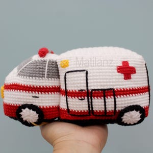 Handmade crocheted ambulance toy in white with red stripes, featuring a gray roof, black wheels with white centers, and a red cross on the side.