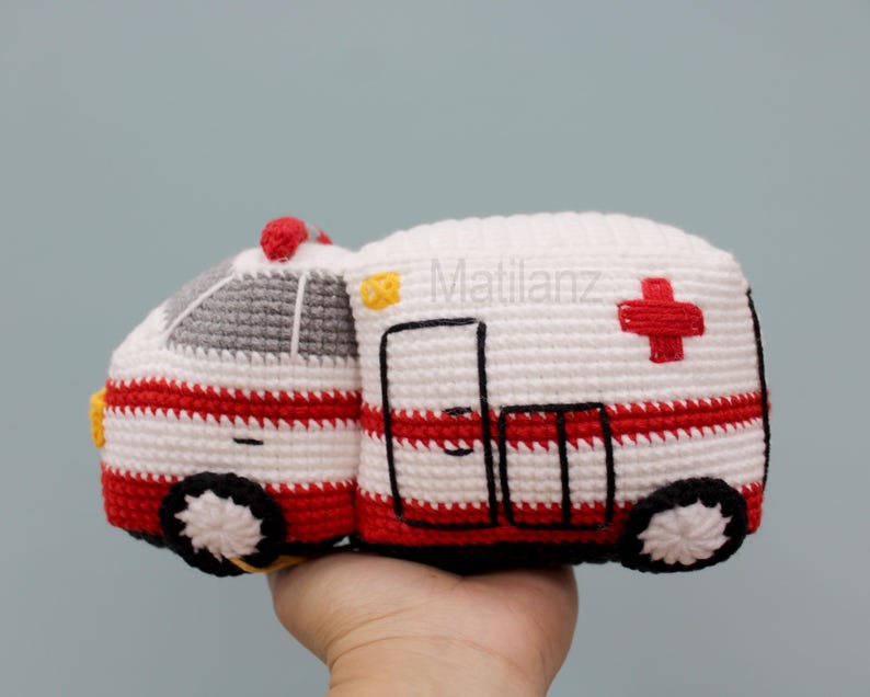 Handmade crocheted ambulance toy in white with red stripes, featuring a gray roof, black wheels with white centers, and a red cross on the side.