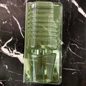 May include: A clear plastic container with two compartments. The top compartment is tall and cylindrical, while the bottom compartment is shorter and wider. The container is made of recyclable PET plastic.