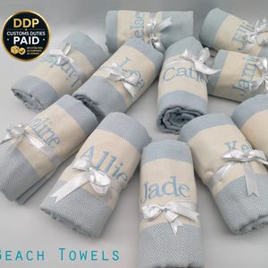 Op de afbeelding: Een verzameling opgerolde strandlakens, elk met een geborduurde naam in lichtblauw. De handdoeken zijn lichtblauw en crème met een visgraatpatroon, vastgebonden met witte satijnen linten. De tekst "BEACH TOWELS" is zichtbaar. Afmetingen: 101,6 x 177,8 cm.