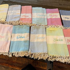 May include: A collection of personalized beach towels in various pastel colors, including pink, blue, yellow, and green. Each towel features a white stripe with an embroidered name and a fringed edge.