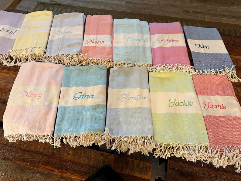 May include: A collection of personalized beach towels in various pastel colors, including pink, blue, yellow, and green. Each towel features a white stripe with an embroidered name and a fringed edge.
