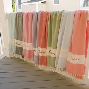 May include: A collection of personalized beach towels in various colors, including green, pink, and coral, hanging on a white railing. Each towel has a white band with a name embroidered in red.