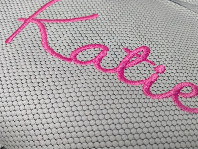 May include: Pink embroidered name "Katie" on a white mesh fabric.