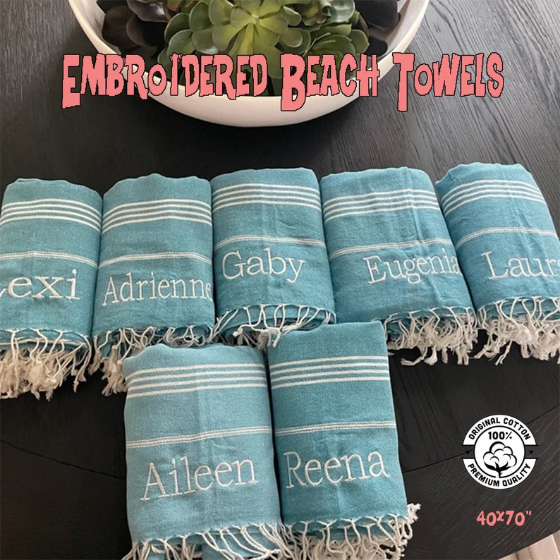 May include: Seven turquoise blue beach towels with white stripes and white fringe. Each towel has a name embroidered on it: Lexi, Adrienne, Gaby, Eugenia, Laur, Aileen, and Reena. The towels are arranged in a semi-circle on a dark wood surface. A white cotton tag with the text "Original Cotton 100% Premium Quality 40x70" is visible in the bottom right corner.