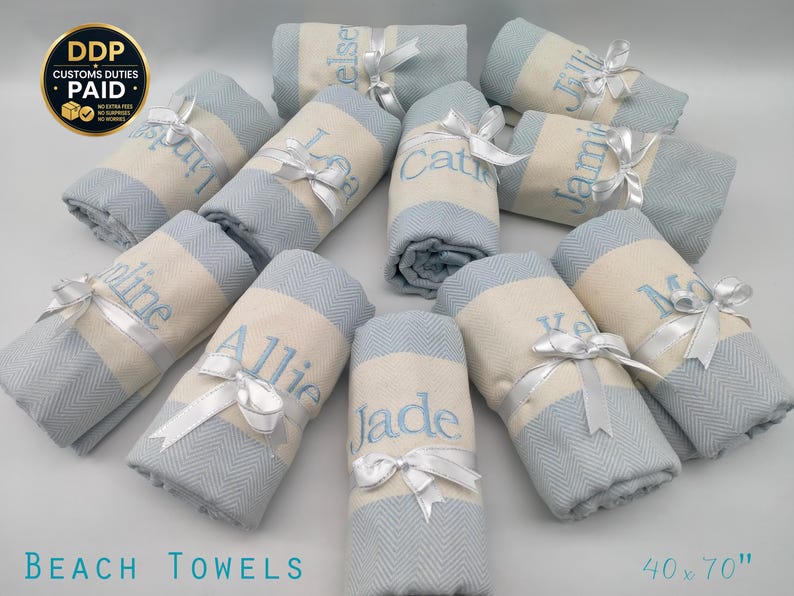 May include: A collection of rolled beach towels, each with a personalized name embroidered in light blue. The towels are light blue and cream with a herringbone pattern, tied with white satin ribbons. The text "BEACH TOWELS" and the dimensions 40 x 70" are visible.