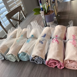 May include: Rolled white towels with embroidered names in various colors, wrapped in sheer white fabric. The towels have patterned edges in shades of blue, pink, and gray. A decorative and personalized gift idea.
