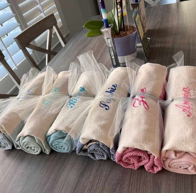 May include: Rolled white towels with embroidered names in various colors, wrapped in sheer white fabric. The towels have patterned edges in shades of blue, pink, and gray. A decorative and personalized gift idea.