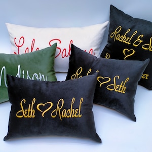 Könnte beinhalten: Sechs dekorative Kissen mit gestickten Namen und einem Herzmotiv. Die Kissen sind in verschiedenen Farben, darunter Schwarz, Weiß, Grün und Gold.