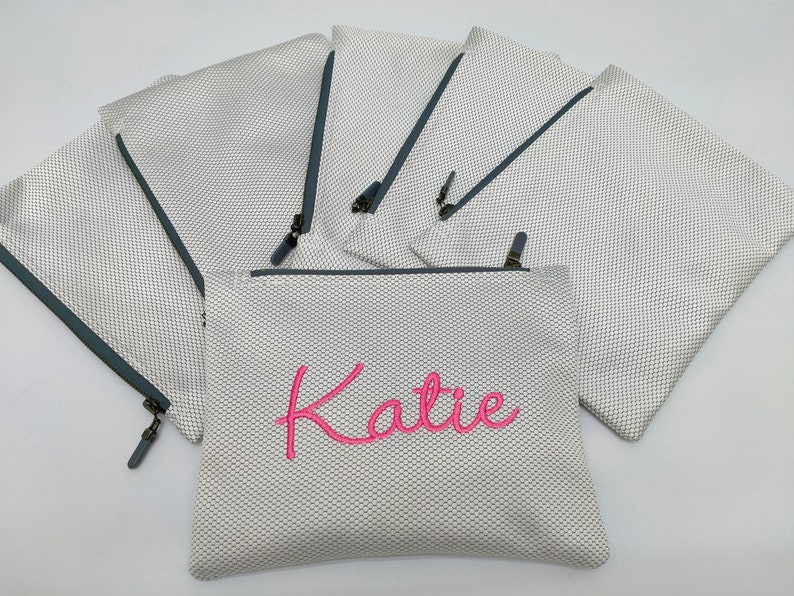 May include: Set of six white mesh zippered pouches with grey trim. One pouch has the name "Katie" embroidered in pink.