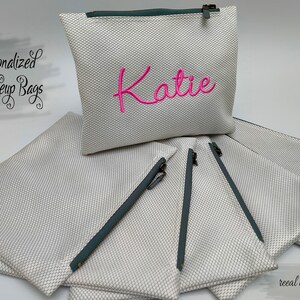 May include: A stack of white zippered makeup bags with grey zippers. The top bag has the name "Katie" embroidered in pink.