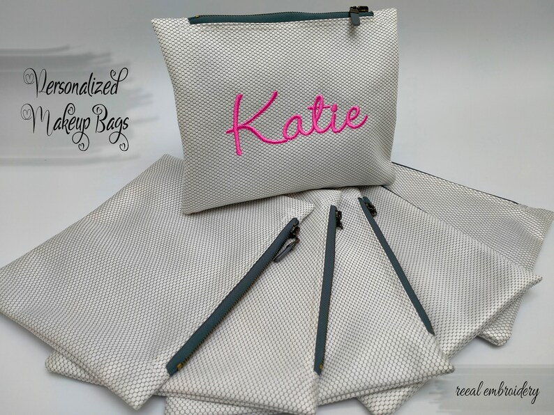 May include: A stack of white zippered makeup bags with grey zippers. The top bag has the name "Katie" embroidered in pink.