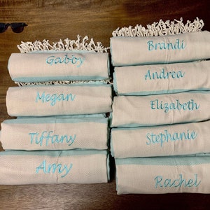 May include: A collection of personalized beach towels with names embroidered in turquoise thread. The towels are rolled up, showing a light blue border and a white, textured body. White fringe decorates the top edge. Sunglasses and a starfish are in the background.