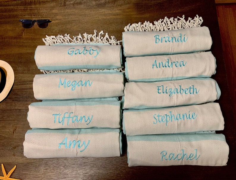 May include: A collection of personalized beach towels with names embroidered in turquoise thread. The towels are rolled up, showing a light blue border and a white, textured body. White fringe decorates the top edge. Sunglasses and a starfish are in the background.