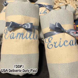 May include: Rolled light blue and cream-colored towels with embroidered names and tied with light blue satin ribbons. The towels have a herringbone pattern on the ends. The names "Camille" and "Erica" are embroidered in blue.