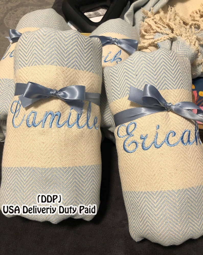 May include: Rolled light blue and cream-colored towels with embroidered names and tied with light blue satin ribbons. The towels have a herringbone pattern on the ends. The names "Camille" and "Erica" are embroidered in blue.