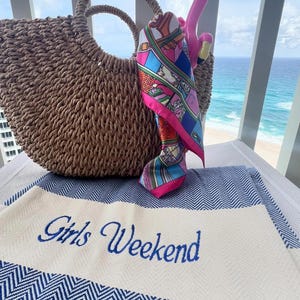 May include: A woven straw tote bag with a round handle, a colorful silk scarf, and a pink flamingo pool float. A blue and white striped beach towel with the words "Girls Weekend" embroidered in blue.