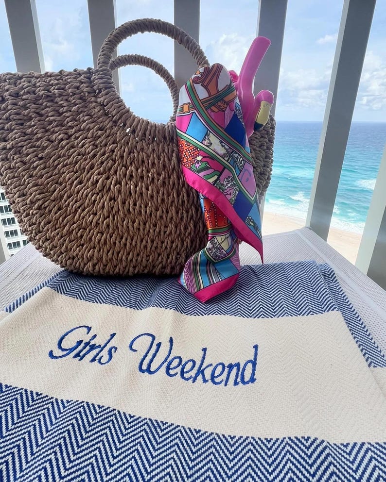 May include: A woven straw tote bag with a round handle, a colorful silk scarf, and a pink flamingo pool float. A blue and white striped beach towel with the words "Girls Weekend" embroidered in blue.