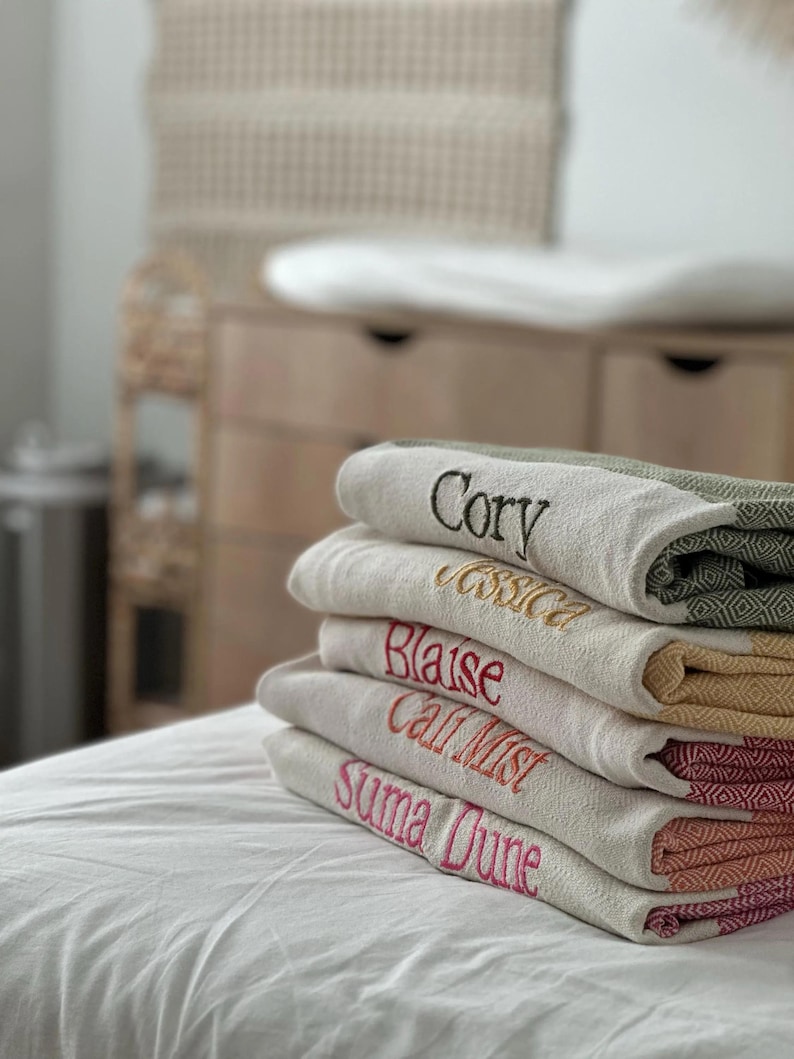 May include: A stack of personalized, embroidered blankets in various colors. Each blanket is a different color and has a name embroidered in a contrasting color. The names include "Cory", "Jessica", "Blaise", "Call Mist", and "Suma Dune".