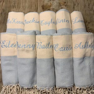 May include: A collection of rolled, personalized towels. Each towel is cream-colored with a light blue chevron pattern on the bottom half. Names are embroidered in light blue thread. The towels have a fringed edge.