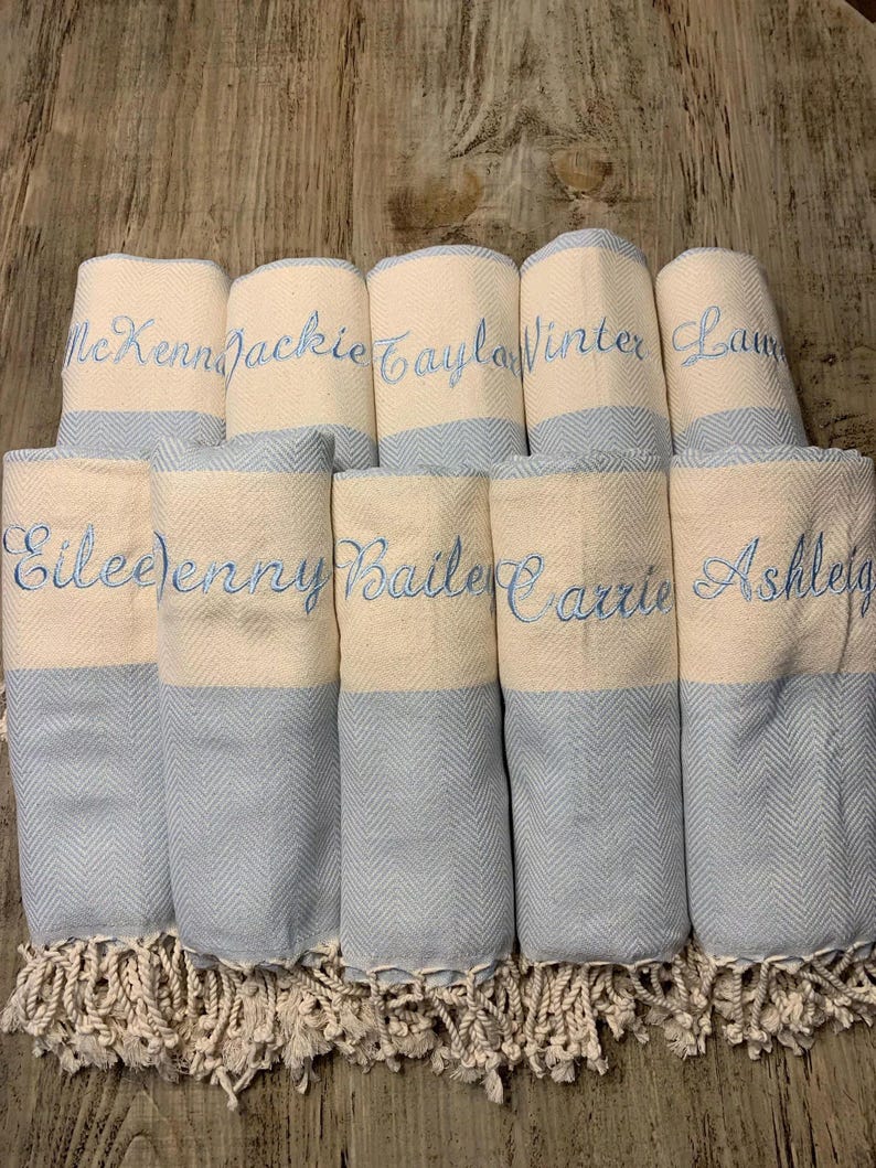 May include: A collection of rolled, personalized towels. Each towel is cream-colored with a light blue chevron pattern on the bottom half. Names are embroidered in light blue thread. The towels have a fringed edge.