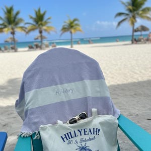 May include: A beach scene with a light blue and white striped towel draped over a chair. The towel is personalized with the name "Hillary". A white tote bag with the text "Hillyeah! 50 & Fabulous" and sunglasses rest on the chair.