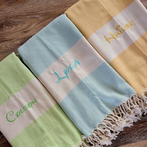 May include: Three personalized beach towels in shades of green, blue, and yellow. Each towel features a cream-colored stripe with embroidered names in coordinating colors. The towels have a diamond pattern and fringed edges.