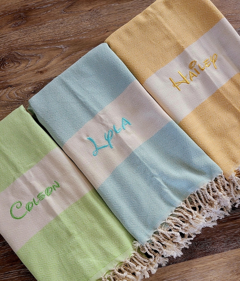 May include: Three personalized beach towels in shades of green, blue, and yellow. Each towel features a cream-colored stripe with embroidered names in coordinating colors. The towels have a diamond pattern and fringed edges.