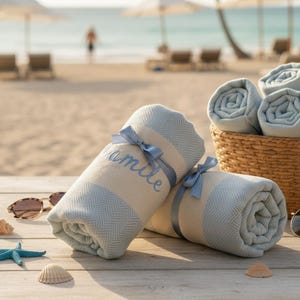 May include: Rolled beach towels with light blue and cream stripes, tied with blue ribbons. One towel has the name "amile" embroidered on it. A basket of rolled towels sits nearby on a wooden table, with sunglasses and seashells.