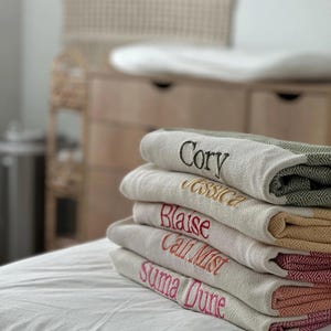 May include: A stack of personalized, embroidered blankets in various colors. Each blanket is a light neutral color with a different name embroidered in a contrasting color. The blankets have a geometric patterned border.