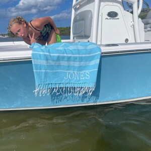 May include: A blue and white striped beach towel with the name "JONES" embroidered on it. The towel is hanging on the side of a white boat.