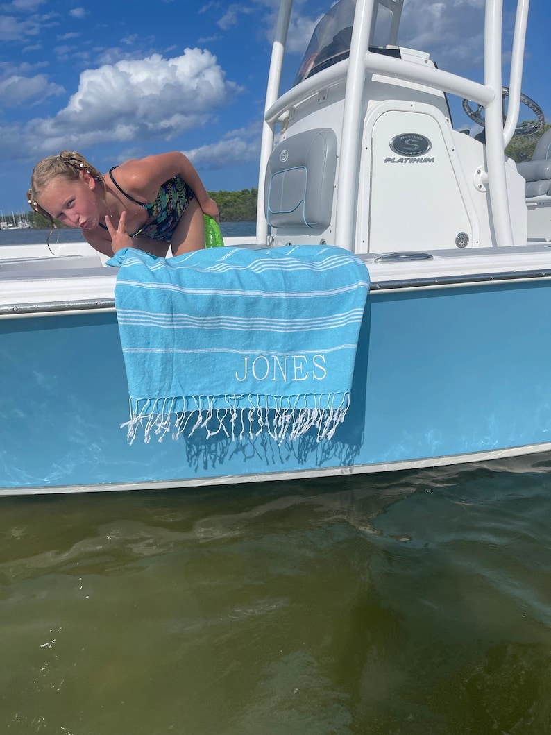 May include: A blue and white striped beach towel with the name "JONES" embroidered on it. The towel is hanging on the side of a white boat.