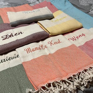 May include: Assortment of personalized Turkish towels in various colors, including green, pink, and blue. Each towel features embroidered names in different fonts and colors. Some towels have geometric patterns and fringe.