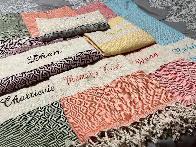 May include: Assortment of personalized Turkish towels in various colors, including green, pink, and blue. Each towel features embroidered names in different fonts and colors. Some towels have geometric patterns and fringe.