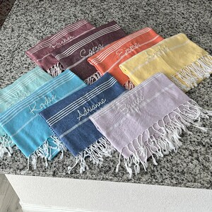 May include: A set of ten personalized towels in various colors, including blue, green, purple, yellow, orange, and red. Each towel has a different name embroidered on it in white thread.