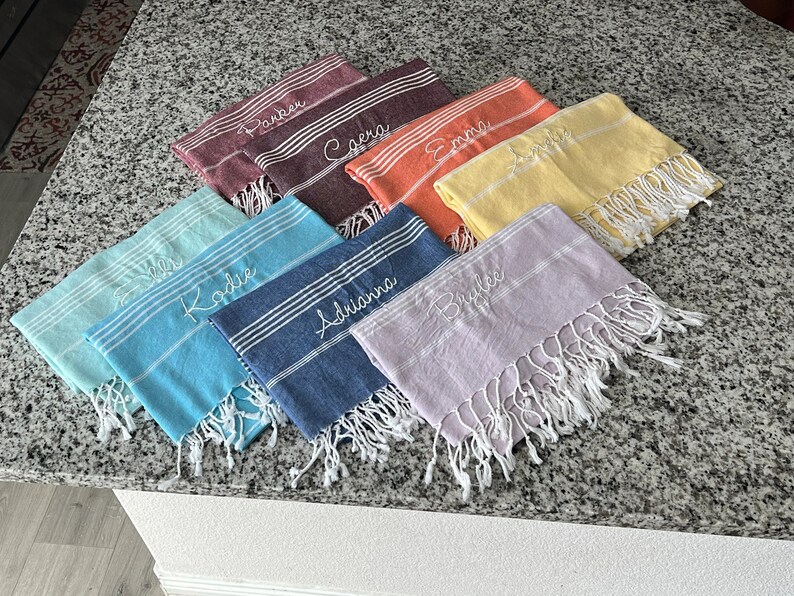 May include: A set of ten personalized towels in various colors, including blue, green, purple, yellow, orange, and red. Each towel has a different name embroidered on it in white thread.